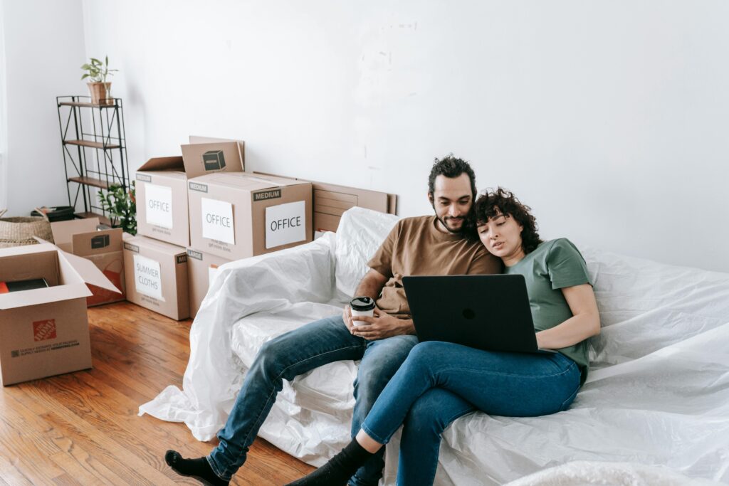 A couple sits together on a couch, using a laptop amidst moving boxes, enjoying relaxed time indoors.